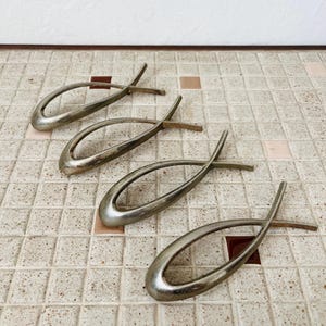 May include: Four silver-toned metal fish-shaped ornaments arranged in a diagonal row. Each piece features a smooth, curved outline forming a stylized fish silhouette with a pointed tail and a rounded head, showcasing a metallic finish with subtle wear.
