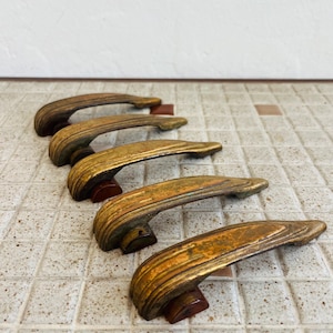 May include: Five curved drawer pulls featuring a metallic gold finish with visible textured ridges and dark brown base accents. Each handle has a tapered, elongated shape with a weathered appearance, arranged in a diagonal row on a tiled surface.
