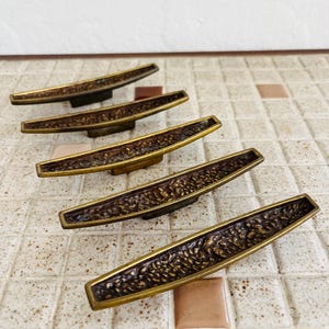 May include: A set of five elongated, curved cabinet pulls made of brass with a dark, textured, and recessed center panel. Each handle features a smooth, polished brass border that frames the intricate, rough, and darkened metal inlay surface.
