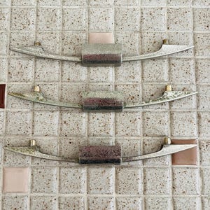 May include: Three silver-toned metal drawer pulls arranged vertically. Each pull features a central rectangular block with two slender, curved arms extending outward, ending in small cylindrical mounting points on the top surface of each arm.
