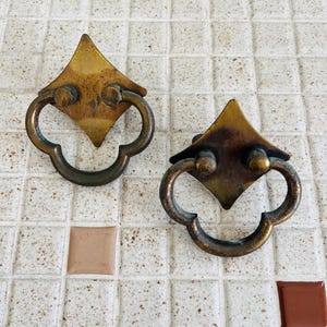 May include: Two brass drawer pulls featuring a diamond-shaped backplate and a rounded, clover-like drop handle. The metal has a weathered, aged patina with dark oxidation spots and a dull, metallic finish on the surface of both hardware pieces.
