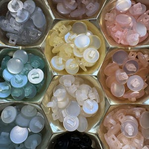 May include: A collection of round, translucent buttons in various colors, including pale pink, light blue, and ivory, are arranged in a hexagonal container. Each button has a shank on the back for sewing. The buttons are made of a shiny, possibly plastic material.