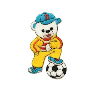 May include: Embroidered patch of a white bear wearing a blue baseball cap with a red "B", a yellow tracksuit, and blue sneakers. The bear is standing on a black and white soccer ball. The patch has a cartoon style.
