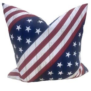 May include: Cushion with a patriotic design. The cushion features a navy blue background with white stars and red and white stripes, resembling the American flag. The stripes have a distressed, wood-like texture.