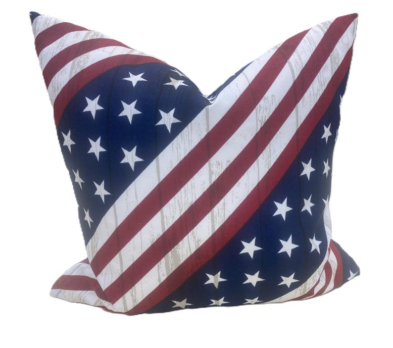 May include: Cushion with a patriotic design. The cushion features a navy blue background with white stars and red and white stripes, resembling the American flag. The stripes have a distressed, wood-like texture.