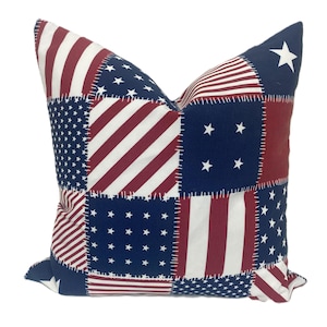 May include: Square decorative pillow with a patchwork design featuring the American flag. The pillow has a navy blue background with white stars and red and white stripes. The seams are stitched with white thread.