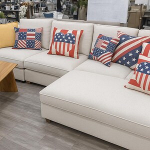 May include: A white corner sofa with cushions featuring American flag patterns in red, white, and blue. A wooden side table with a metal tub is in the foreground. The sofa is a prominent piece of furniture.