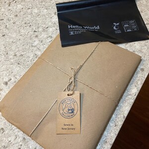 May include: A parcel wrapped in brown paper, secured with twine, and a tag reading "Softness Tailoring, Sewn in New Jersey." A black compostable bag with "Hello World" is on top.