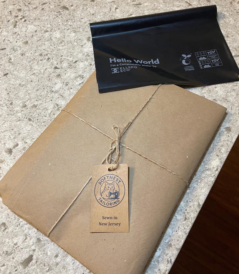 May include: A parcel wrapped in brown paper, secured with twine, and a tag reading "Softness Tailoring, Sewn in New Jersey." A black compostable bag with "Hello World" is on top.