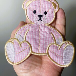 Op de afbeelding: Een roze en witte gingham teddybeer applique met gouden stiksels. De beer zit met zijn poten voor zich.