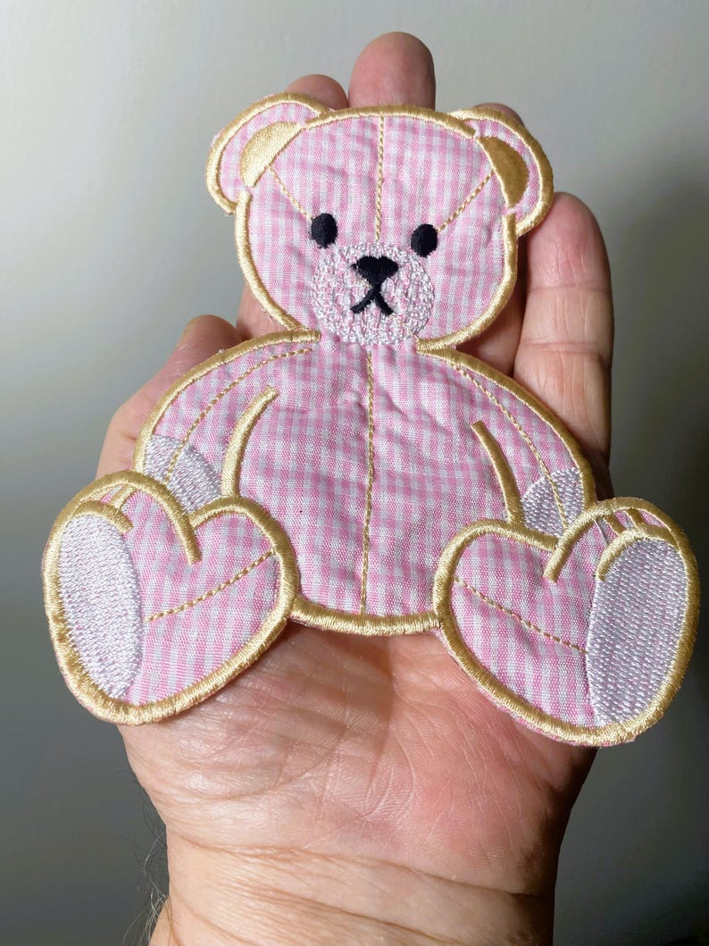Op de afbeelding: Een roze en witte gingham teddybeer applique met gouden stiksels. De beer zit met zijn poten voor zich.