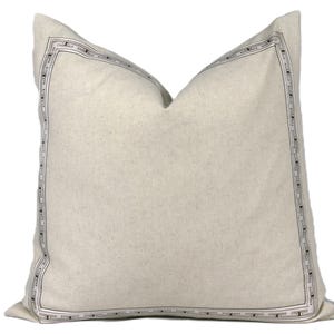 May include: A square, off-white decorative pillow with a geometric border design in black and silver. The pillow has a soft, textured appearance and is likely made of linen or a similar fabric. The border frames the pillow's edges.