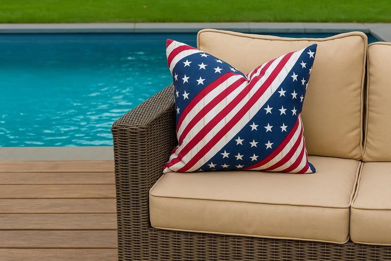 May include: A cushion with a design inspired by the American flag. The cushion has a blue field with white stars and red and white stripes. It sits on a beige outdoor sofa with a wicker frame, near a swimming pool.