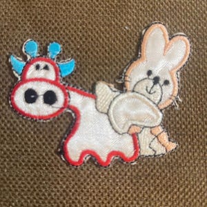 May include: A white felt applique of a rabbit riding a red and white cow with blue horns. The rabbit has a pink nose and brown eyes. The cow has black eyes.