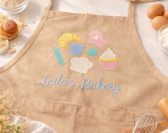 May include: Tan apron with embroidered baking-themed designs, including a croissant, cupcake, and chef's hat. The text "Laila's Bakery" is embroidered in blue script. The apron has a front pocket and adjustable neck strap.