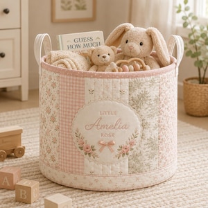 Puede incluir: Una cesta de almacenamiento cilíndrica de tela con un diseño de patchwork acolchado en tonos rosa, crema y verde salvia. El frente muestra un parche festoneado bordado con el texto "LITTLE Amelia ROSE" y un pequeño lazo de cinta rosa.