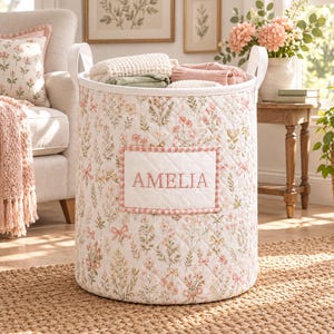 May include: A cylindrical quilted fabric laundry basket featuring a white base with a repeating pattern of pink flowers, green foliage, and small bows. A rectangular white patch on the front is embroidered with the name AMELIA in pink thread.