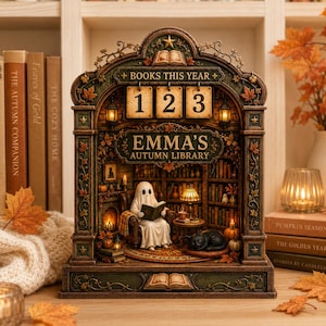 May include: A decorative tabletop calendar featuring an arched library scene with a ghost reading in a chair, a black cat, and shelves of books. The text reads BOOKS THIS YEAR and EMMA'S AUTUMN LIBRARY. The structure is adorned with autumn leaves.