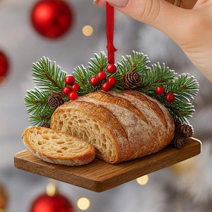 May include: A Christmas ornament shaped like a loaf of bread with a slice cut off, resting on a wooden cutting board. The ornament is decorated with pine needles, red berries, and small pine cones. A red ribbon is attached for hanging.