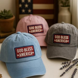 Pode incluir: Três bonés de beisebol em azul, rosa e cinza, cada um com um patch retangular que diz "GOD BLESS AMERICA" em texto branco com uma estrela vermelha. Os bonés são exibidos em uma superfície branca com uma bandeira americana emoldurada ao fundo.