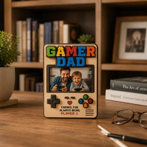 Könnte beinhalten: Ein Holzfotorahmen mit den Worten "GAMER DAD" in bunten Buchstaben. Der Rahmen ist wie ein Videospiel-Controller gestaltet, mit einem Foto von Vater und Sohn beim Videospielen. Der Text "THANKS FOR ALWAYS BEING PLAYER 1" ist ebenfalls enthalten.