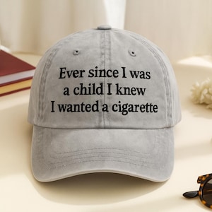 May include: Gray baseball cap with the text "Ever since I was a child I knew I wanted a cigarette" in black. The hat is sitting on a table with a book, flowers, and sunglasses.