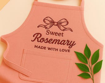 May include: A light pink apron with a pocket and a neck strap. The apron has the words "Sweet Rosemary MADE WITH LOVE" embroidered in a dark red color, along with a bow design. A sprig of green leaves is placed on the right side.