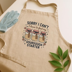 May include: Beige linen apron with the text "SORRY I CAN'T I have to feed my SOURDOUGH STARTER." The apron features a graphic of three jars with pink lids and tags. A pair of light blue gloves are on the left.