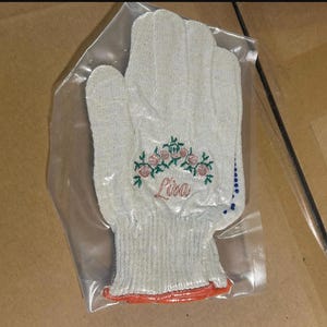 May include: A light gray work glove with an embroidered floral design and the name "Lisa" in cursive. The glove is in a clear plastic bag. The cuff has an orange trim.