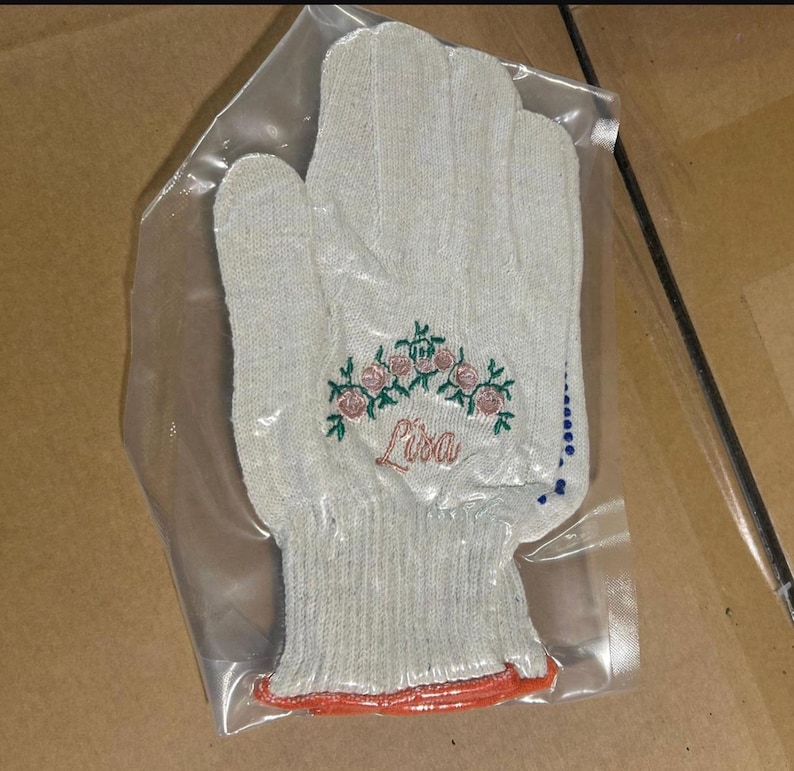 May include: A light gray work glove with an embroidered floral design and the name "Lisa" in cursive. The glove is in a clear plastic bag. The cuff has an orange trim.