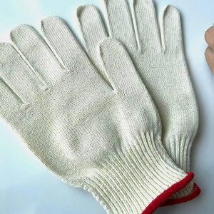 May include: A pair of off-white work gloves with a red trim on the cuffs. The gloves are made of a knitted material and are designed to protect the hands. The gloves are shown on a white surface.