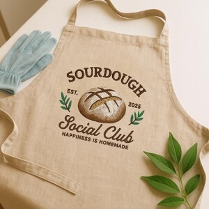 Pode incluir: Avental de linho bege com o texto "SOURDOUGH Social Club" e um gráfico de um pão. O avental também apresenta o texto "EST. 2025" e "HAPPINESS IS HOMEMADE". Um par de luvas azul claro está no canto superior esquerdo.