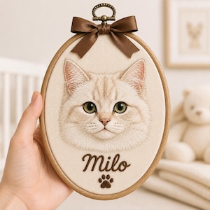 Puede incluir: Un bastidor de bordado ovalado de madera que contiene un panel de tela color crema con un retrato bordado detallado de un gato claro con ojos verdes. Debajo del gato, el nombre Milo y una huella están bordados en marrón. Un lazo marrón corona la parte superior.