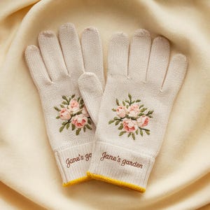 May include: A pair of light beige knitted gardening gloves. Each glove features an embroidered floral design with pink roses and green leaves. The cuffs have the text "Jane's garden" and a yellow trim.