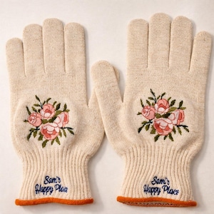 May include: A pair of beige knit gloves with embroidered pink roses and green leaves. The gloves have an orange trim and the words "Sam's Happy Place" in blue script. The gloves are designed for warmth and style.