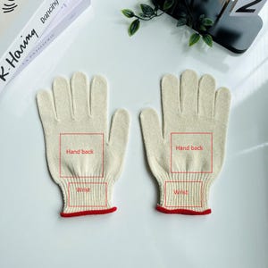 May include: Two off-white knitted gloves with red trim at the wrist. The gloves are laid out on a white surface. The words "Hand back" and "Wrist" are written on the gloves.