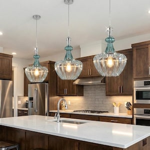 May include: Three pendant lights with clear, ribbed glass shades and teal accents hang above a kitchen island. The lights have a vintage-inspired design with a silver finish and are suspended from the ceiling.
