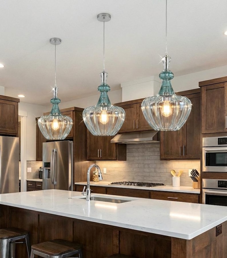 May include: Three pendant lights with clear, ribbed glass shades and teal accents hang above a kitchen island. The lights have a vintage-inspired design with a silver finish and are suspended from the ceiling.