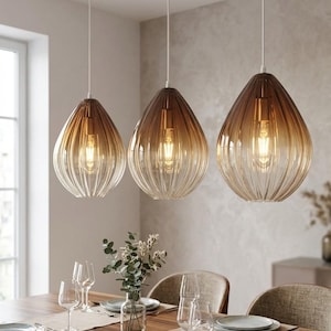 Op de afbeelding: Drie druppelvormige hanglampen met geribbelde glazen kappen met een bruin-naar-transparante gradiënt. Elke lamp hangt aan een wit snoer en bevat een zichtbare gloeiende lamp, opgehangen boven een houten eettafel.