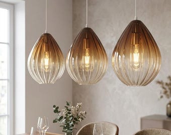 May include: Three teardrop-shaped pendant lights with ribbed glass shades featuring a brown-to-clear gradient finish. Each light hangs from a white cord and contains a visible glowing light bulb, suspended above a wooden dining table.