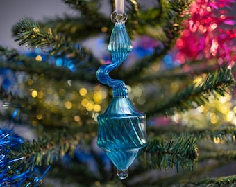 Puede incluir: Un adorno navideño de cristal turquesa con un diseño en espiral cuelga de una cinta blanca. El adorno está frente a un árbol de Navidad decorado con oropel y luces verdes, azules y rosas.