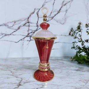 Könnte beinhalten: Eine dekorative rote Glasparfümflasche mit goldenen Akzenten. Die Flasche hat eine konische Form mit rundem Boden und einem mattierten weißen Deckel. Der Deckel ist mit goldenen Zierleisten und einem transparenten Ringgriff verziert. Die Flasche ist etwa 20 cm hoch.
