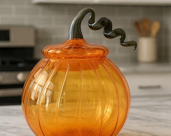 May include: An orange glass pumpkin-shaped jar with a lid and a dark green, swirled stem. The jar has vertical ridges and sits on a marble countertop. The background includes a kitchen with white cabinets and a gray backsplash.