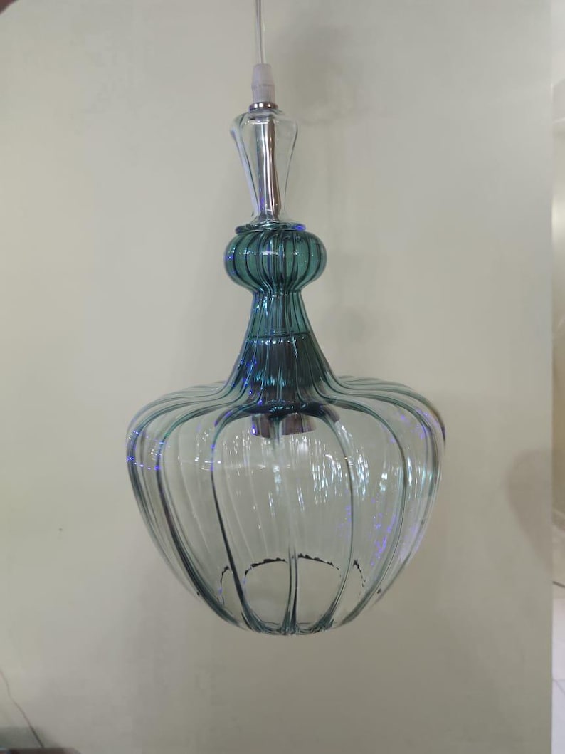 May include: A clear glass pendant light with a teal-colored top and a bulbous, ribbed body. The light fixture hangs from a white cord and features a decorative, elongated top.