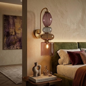 May include: A modern wall sconce with a stacked glass design in shades of purple, gray, and brown. The fixture has a brass arm and base, and a single light bulb. The sconce is mounted on a beige wall near a bed.