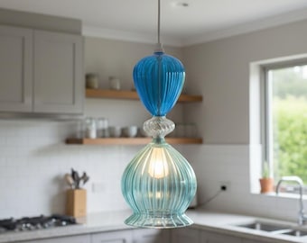 May include: A hanging pendant light with a two-tiered, blue and turquoise glass shade. The light fixture has a ribbed design and hangs from a white cord. The light is turned on, illuminating the glass.