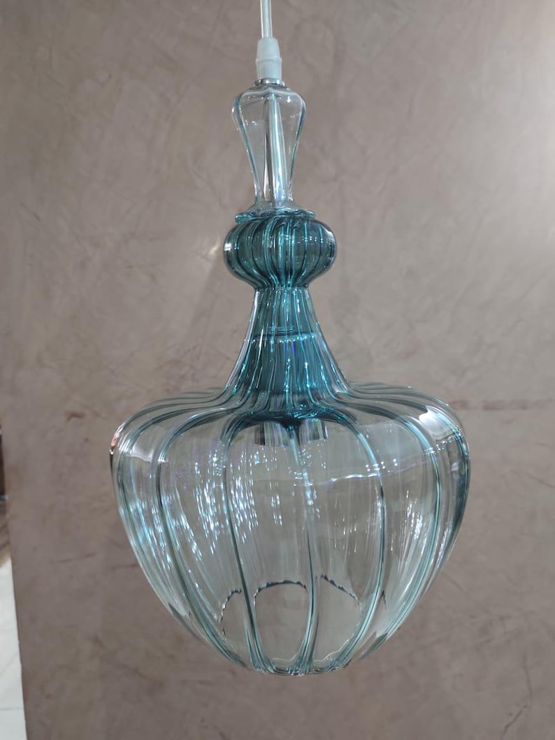 May include: A clear, teal-colored glass pendant light fixture. The light has a bulbous, ribbed shade and a slender, decorative top. The fixture is suspended by a white cord, showcasing a vintage or art deco design.