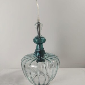 May include: A clear glass pendant light with a teal-colored top and a ribbed, rounded body. The light fixture hangs from a clear cord and has a silver-colored base.