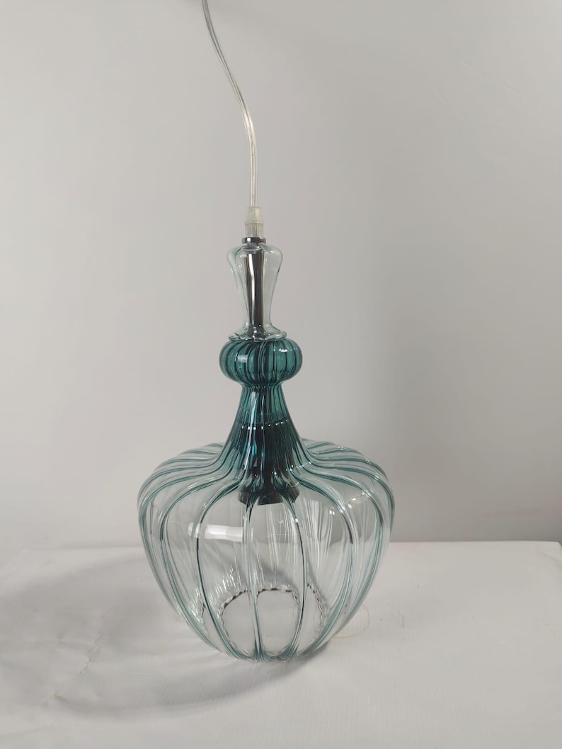 May include: A clear glass pendant light with a teal-colored top and a ribbed, rounded body. The light fixture hangs from a clear cord and has a silver-colored base.