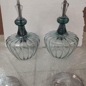May include: Two glass pendant lights with a teal green top and a clear, ribbed glass body. The lights have a decorative shape and are suspended by a silver cord. Two silver ceiling mounts are in the foreground.
